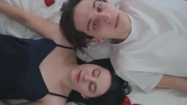 Sevgi dolu genç bir çift beyaz yatakta uzanmış gülümsüyor ve üst manzarayı okşuyor. Evde dinlenen mutlu beyaz erkek ve kadının yakın plan portresi.