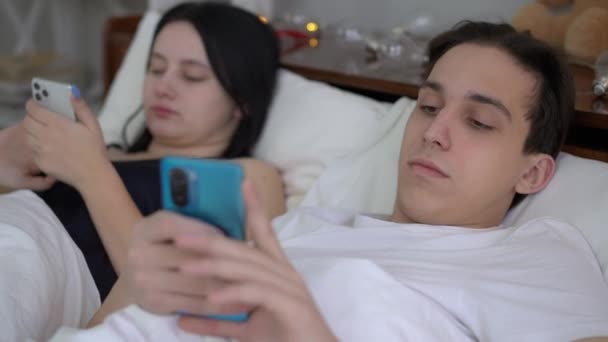 背景にベッドに横たわってオンラインでぼやけたガールフレンドメッセージングとしてスマートフォンアプリをスクロール若いボーイフレンドの肖像画 家の中でベッドルームで不注意な白人カップル — ストック動画