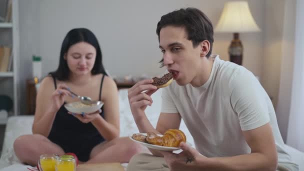 リラックスした白人男性の肖像は背景にベッドの上に座って手を横断オートミールと女性を不承認として午前中においしい甘いデザートを食べています ライフスタイルと栄養の概念 — ストック動画