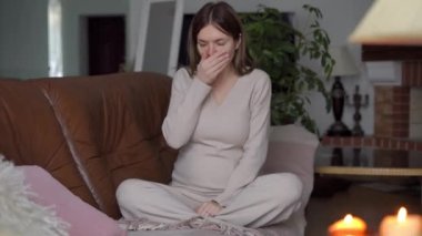 Oturma odasındaki kanepede oturmuş öksüren hamile bir kadın. Soğuk algınlığı ve grip virüsü belirtileri gösteren üzgün, hasta, beyaz bir kadının geniş açılı portresi.