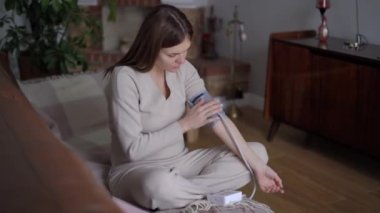 Oturma odasındaki koltukta oturan ve kan basıncını ölçen üzgün, hasta, genç bir kadın. Kapalı alanda sfenometre ile kafkasyalı beklenenin geniş açılı portresi.