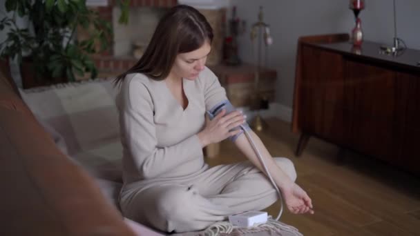 若い悲しい病気の妊婦は リビングルームのソファに座っているデバイスをオンに血圧を測定します Wide Shot Portrait Unwell Upset Caucasian Expectant Sphygmomanometer — ストック動画