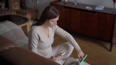 Geniş açılı görüntü, genç hamile bir kadının dijital tablet dokunmatik ekranda telefon konuşması yapması. Kendine güveni tam, beyaz, oturma odasında kanepede oturuyor.