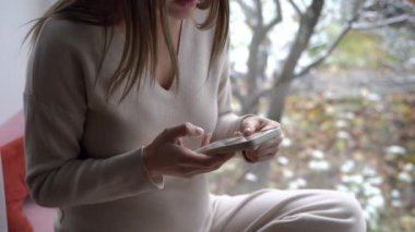 Tanımlanamayan hamile bir kadın pencerenin kenarında oturmuş sesli mesaj kaydediyor. Beyaz adam evde akıllı telefon uygulaması kullanıyor.