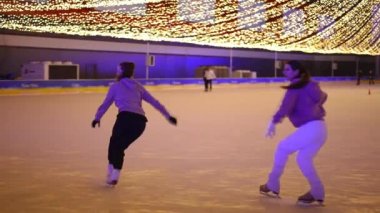 Ağır çekimde buz patenleriyle zıplayan iki sporcu kadın tek ayak üstünde yürüyor. Canlı kamera, kendine güveni tam beyaz kadınları buz pateni sahasında antrenman yaparken izliyor.