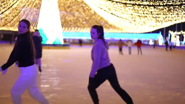 屋外のアイスリンクでゆっくりと動きながらアイススケートに乗る2人の優雅な若い白人女性 自信を持ってスリム美しいスポーツ女性のトレーニングフィギュアスケート夜の街で — ストック動画