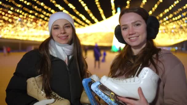 スローモーションで笑顔カメラを見てアイススケートを持つ2人の若い幸せなスポーツ女性 自信に満ちた白人女性の肖像画でクリスマスアイスリンクで夜市 — ストック動画