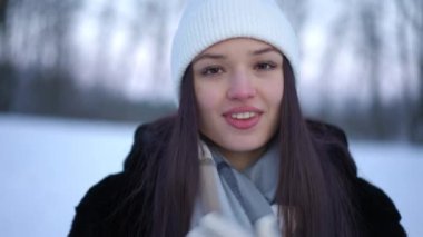 Yavaş çekimde şapkaya dokunan, kahverengi gözlü ve dişlek gülüşlü mutlu genç kadın portresi. Sabah kış ormanlarında poz veren büyüleyici beyaz kadın.