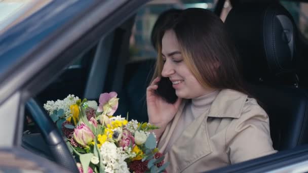 正面年轻女子笑着在电话上与花在车上交谈的侧视图肖像 快乐的白人女士在外面的汽车花束 — 图库视频影像