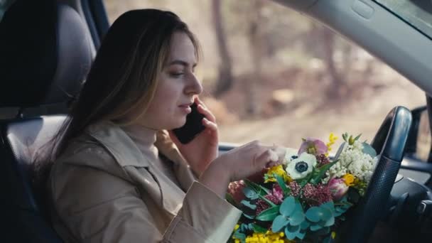 年轻女人在电话里笑着坐在外面的车上 开着鲜花 车载着花束在智能手机上聊天的迷人的白人女士的侧视图画像 — 图库视频影像