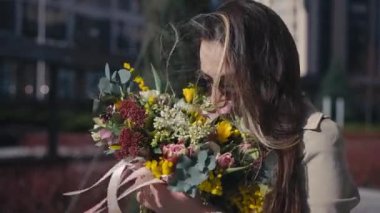 Güneş gözlüklü esmer bir kadın, gülümsüyor ve şehir caddesinde güneşin altında duruyor. Açık havada çiçekli zarif, kendine güvenen beyaz kadın portresi.