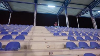 Geniş açılı stadyum tribünleri, üstünde bulanık beyaz kadın oturuyor. Tanımlanamayan bir bayan geceleri spor sandalyesinde düşünmekten zevk alıyor.