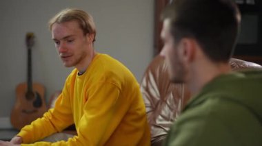 Oturma odasındaki kanepede oturan genç, pozitif sarışın bir adam arkadaşıyla konuşuyor. Rahatlamış beyaz adamın portresi evde arkadaşla eğleniyor.