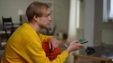 Basketbol topu gibi uzaktan kumandayla kanal değiştiren genç bir adam. Oturma odasında televizyon izleyen konsantre beyaz adamın portresi.