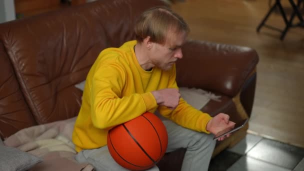 Ξανθός Καυκάσιος Νεαρός Κάθεται Στον Καναπέ Μπάλα Μπάσκετ Βλέποντας Οθόνη — Αρχείο Βίντεο