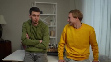 Masaya yaslanmış gülümseyen iki rahat genç adam konuşuyor. Kendine güvenen beyaz erkek arkadaşlarının portresini büyüt. Hafta sonları oturma odasında sohbetin tadını çıkar.
