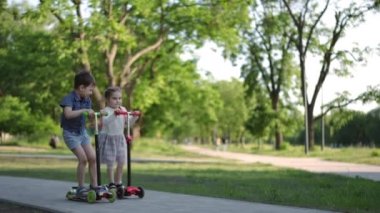 Geniş açılı mutlu küçük çocuk ve kız ağır çekimde hareket eden scooterlara biniyor. Heyecanlı beyaz çocuklar yaz parkında eğleniyorlar.