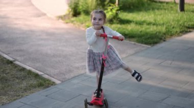 Yüksek açılı, kaygısız küçük kız scooter 'da dengede duruyor bir bacağını yavaş çekimde yana açıyor. Kendine güvenen beyaz çocuğun geniş açılı portresi park alanında eğleniyor.