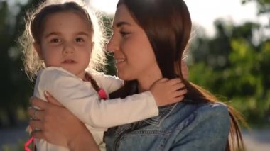 Güneşin altında duran çocukla konuşan küçük kızı öpen mutlu genç kadının yakın plan portresi. Kendine güvenen güzel beyaz anne yaz parkında gün batımında kızıyla eğleniyor.