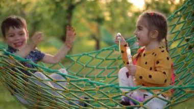 Yan bakışta gülen rahat çocuklar yaz parkında hamakta oturup sabun köpüğü yakalıyorlar. Kaygısız Kafkasyalı erkek ve kız gülerek gün batımının tadını çıkarıyorlar.