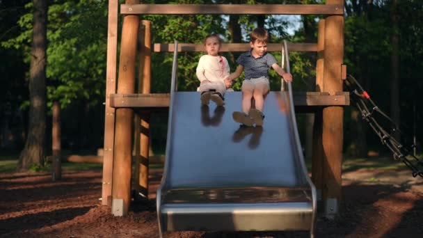リラックスした男の子と女の子が上に座って お互いの笑顔を見つめながら 子供の遊び場でワイドショットスライド スローモーションで公園で夏のレジャーを楽しんでいるハッピーな白人友人 — ストック動画