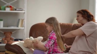 Yandan bakıldığında, oyuncak ayıyla oynayan rahat bir kız uzun sarı saçları tarayan bir kadındır. Oyuncağı ve annesi olan mutlu beyaz kız evde çocuğuna bakıyor.
