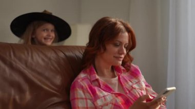 Konsantre kadın akıllı telefonların dokunduğu ekranda Cadılar Bayramı cadı kostümlü neşeli kız olarak elleri ile gözlerini kapatıyor. Mutlu Kafkas anne ve kızı içeride eğleniyor.