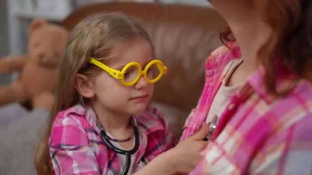 迷人的快乐女孩戴着黄色的玩具眼镜 听着听不清的福音书 像是一个难以辨认的快乐女人咳嗽着 白人阳性女儿检查母亲模拟医生在室内玩耍 — 图库视频影像