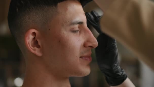 中东青年男子侧视脸 白种女人给男性眉毛打蜡 室内美容温泉疗程中带着微笑自信的病人的特写 — 图库视频影像