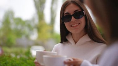 Kendine güveni tam genç bir kadının portresi kahve içerken açık kafede arkadaşlarıyla otururken sağdan sola doğru çekilen canlı kamera görüntüsü. Gülümseyen beyaz kadın, şehir merkezinde sıcak içkinin tadını çıkarıyor.