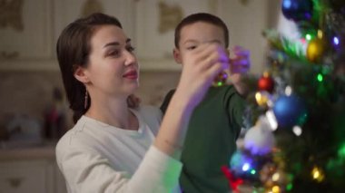 Oğlu kollarında bir Noel ağacına top asan bir annenin yakın çekimi. Çocuk, annesi Noel ağacına bir Noel ağacı oyuncağı asmasına izin vermediği için üzgündü. Gülümse anne.