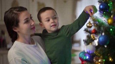Annesinin kollarında mutlu bir çocuk Noel ağacına bir Noel topu asıyor. Mutlu anne ve oğul Noel partisine hazırlanıyor.