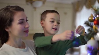 Annesinin kollarında gülümseyen bir çocuğun Noel ağacına mavi bir Noel topu asışının yakın çekimi. Noel arifesinde mutlu anlar