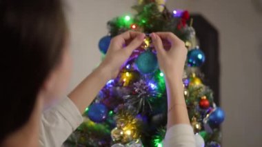 Bir kadının iki eliyle bir Noel ağacına astığı turkuaz Noel ağacı topunun omzunun üstünden.