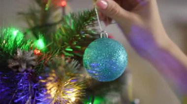 Bir kadının Noel ağacının dalına asıldığı turkuaz bir Noel topunun yakın çekimi. Sağ eli ağır çekimde.