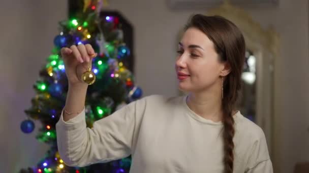 クリスマスツリーの背景に対して 彼女が手に持っている黄色く輝くクリスマスボールを見ている美しい女の子のクローズアップ — ストック動画