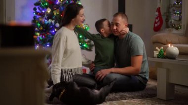 Ağır çekim. Mutlu bir aile, odadaki yerde bir Noel ağacının yanında oturuyor. Oğlu da karşılığında ailesini öper, onlar da onu öper. Yeni yıl ve Noel konsepti. Mutlu aile kutlaması