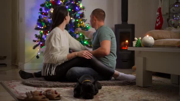 スローモーション 美しい結婚したカップルは 暖炉のカーペットの上に座って クリスマスツリーに掛かっている青いクリスマスボールを元気に調整します — ストック動画