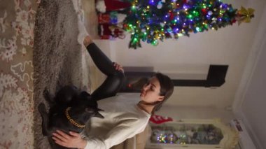 Dikey video. Yavaş çekim. Oturma odasında halıya oturmuş bir kız Noel ağacının arka planında küçük siyah bir köpeği okşuyor.