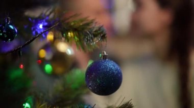 Ağır çekim. Noel ağacı dalında mavi bir Noel balosunun yakın çekimi. Arka planda, öpüşen bir çift odak dışı.