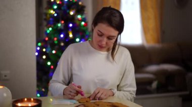 Ağır çekim. Noel için dekore edilmiş modern bir evde masada oturan güzel bir kadın Noel kurabiyesi süslüyor.