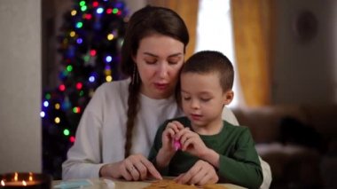 Ağır çekim. Evde, Noel ağacının arka planında masada oturan anne, oğlunun Noel kurabiyelerini yenilebilir şekerli kalemlerle süslemesine yardım ediyor. Hazırlıklar