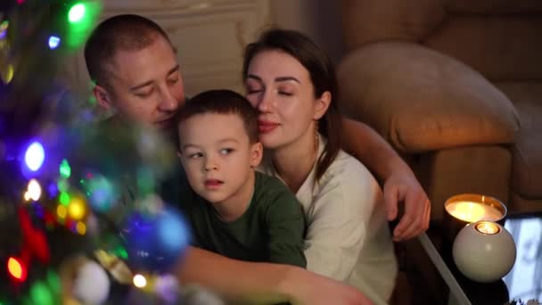 スローモーション クリスマスツリーの近くの家のクリスマスに強い家族が抱擁します 男は妻と息子を両手で抱きしめている 男の子が両親にキスをする クリスマスと新年の家族のレジャー時間 — ストック動画