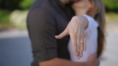 Bir kadın, bir erkeğin kollarında, kameraya evlilik yüzüğüyle elini gösterir. Öpüşen çift odak dışı