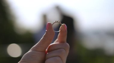 Yakın plan. Bir kadın, iki parmağıyla tuttuğu nişan yüzüğüne bir bahar parkındaki bir çiftin arka planına bakıyor. Evlilik teklifi.