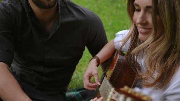 一个快乐的女人坐在城市公园的草地上 为她的情人弹奏吉他 — 图库视频影像