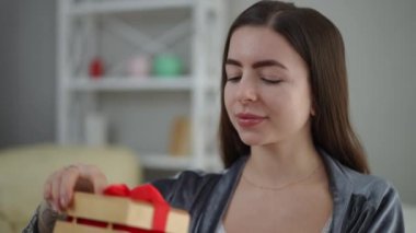 Yakın plan. Kız elinde kırmızı kurdeleyle bağlanmış güzel bir ahşap hediye kutusu tutuyor. Kutuyu açıp hediyeyi görünce, kız sevinçle gülümsüyor.