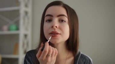 Yakın plan. Kameraya bakarken, uzun saçlı güzel bir kadın dudaklarını dudak parlatıcısıyla boyuyor. Kız modern bir evin yatak odasında makyaj yaparken gülümsüyor.