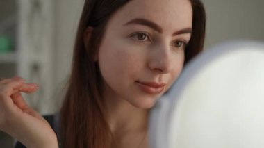 Elindeki aynaya bakan uzun saçlı güzel bir kadın sabah makyajını olumlu değerlendiriyor. Kız gülümsüyor, aynada kendine bakıyor, yatak odasında oturuyor. Yakın plan.