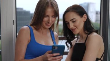 İki güzel beyaz kız bir kadının elindeki cep telefonunun ekranına bakıyor. Modern bir apartman dairesinde açık bir pencerenin önünde duran kızlar konuşup gülüyor.